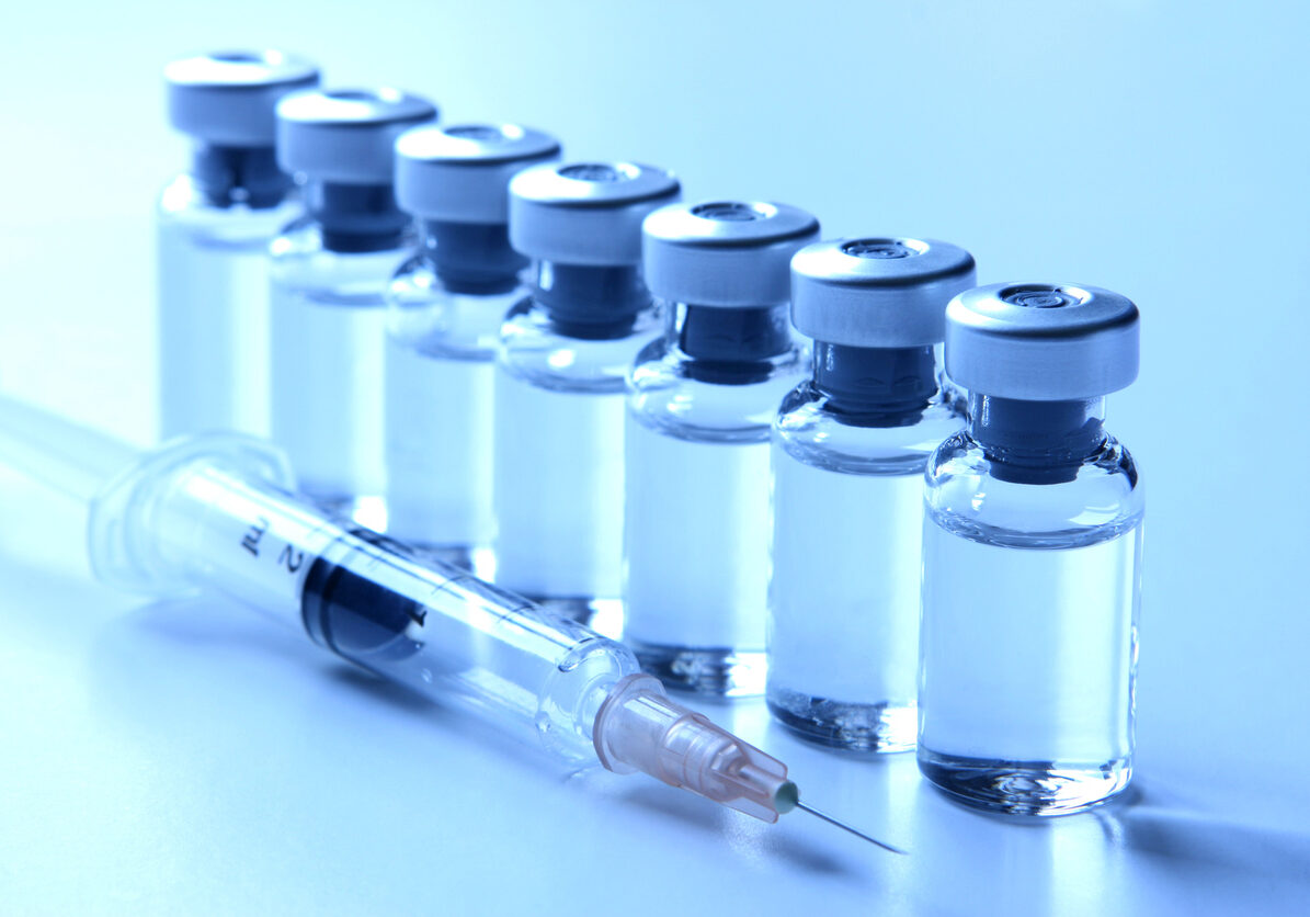 Studio shot of a series of seven vials standing in a row filled with liquid medication. Empty syringe laying next to the vials is ready for injection and  could represent concepts like vaccination, flu shot, medication. Image is blue tinted.